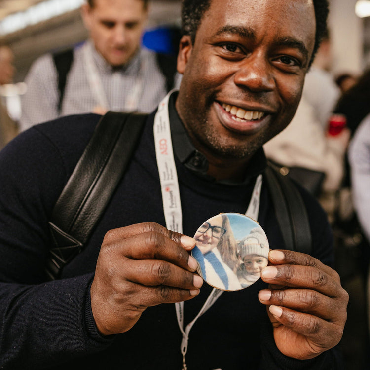A smiling man in a dark sweater holds up a round custom photo-printed biscuit featuring a couple, with a lanyard around his neck and an indoor event background.