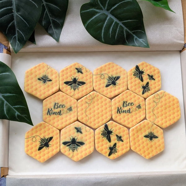 A set of yellow honeycomb-patterned hexagonal iced biscuits with black bee illustrations and the phrase 'Bee Kind' printed on two of them, displayed in an open box with green leaves around them.