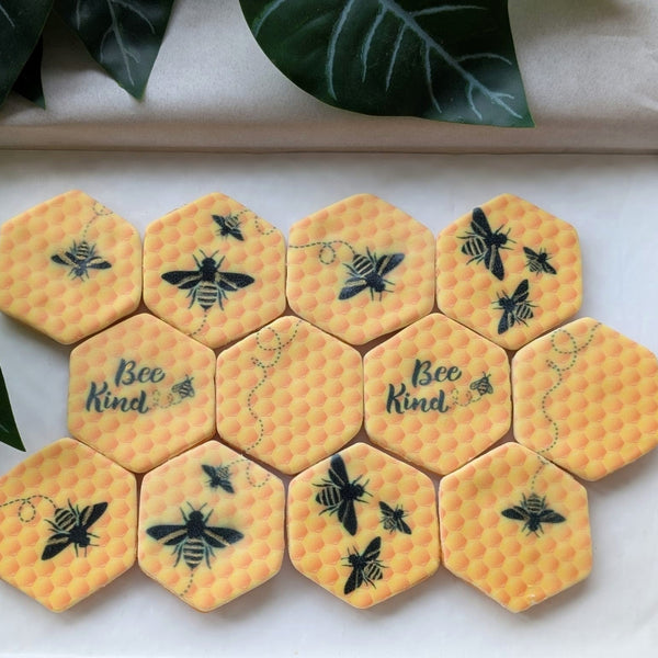 A set of yellow hexagonal beeswax-style fondant biscuits with black printed bee designs and two reading 'Bee Kind,' arranged in a letterbox-friendly pack with decorative green leaves around them.
