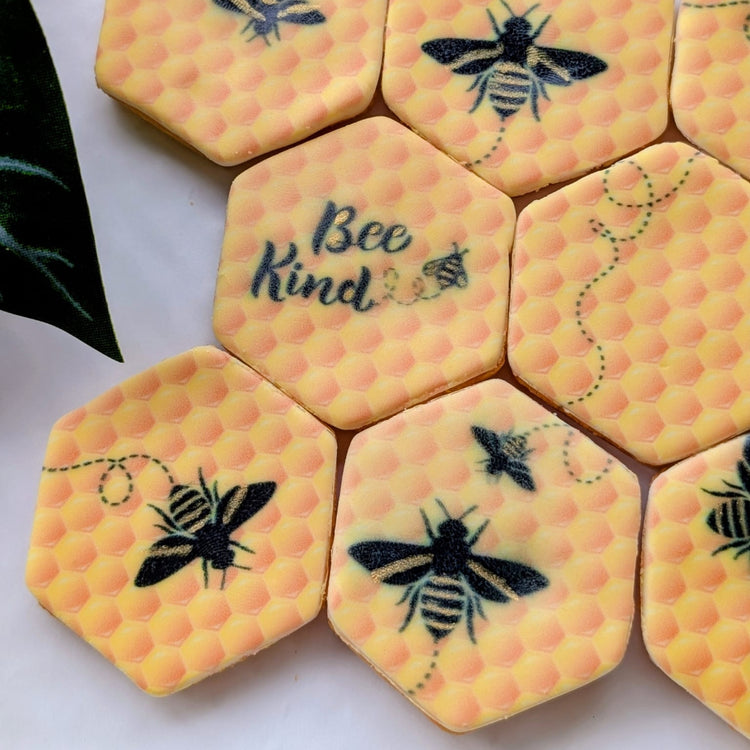 Hexagonal yellow cookies decorated with black bumblebee illustrations and gold detailing. One cookie featuring the words 'Bee Kind' in script, arranged on a white surface.
