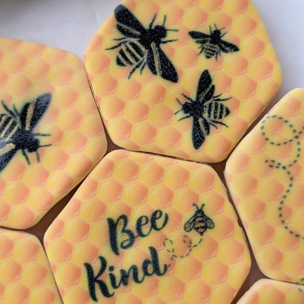 Hexagonal yellow honeycomb-patterned fondant cookies with black bee illustrations, gold detailing and one reading 'Bee Kind' on a white surface.