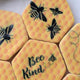 Hexagonal yellow honeycomb-patterned fondant cookies with black bee illustrations, gold detailing and one reading 'Bee Kind' on a white surface.