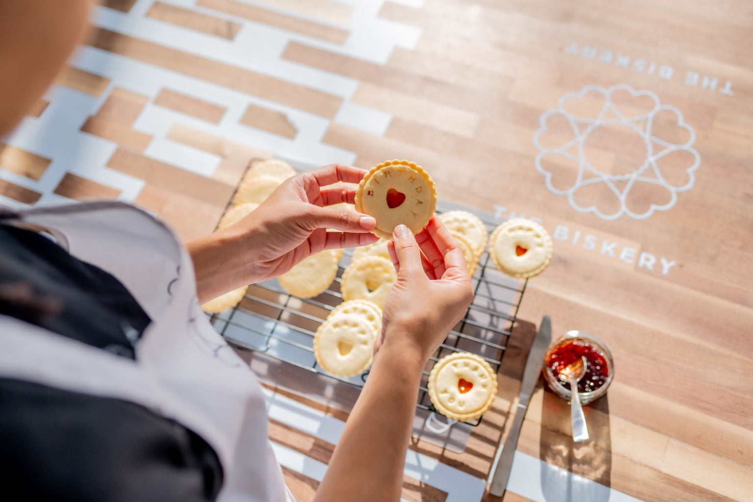 Baker hold jam biscuit impressed with the word birthday. against backdrop of biscuits cooling on a rack, jam and branded The Biskery table.