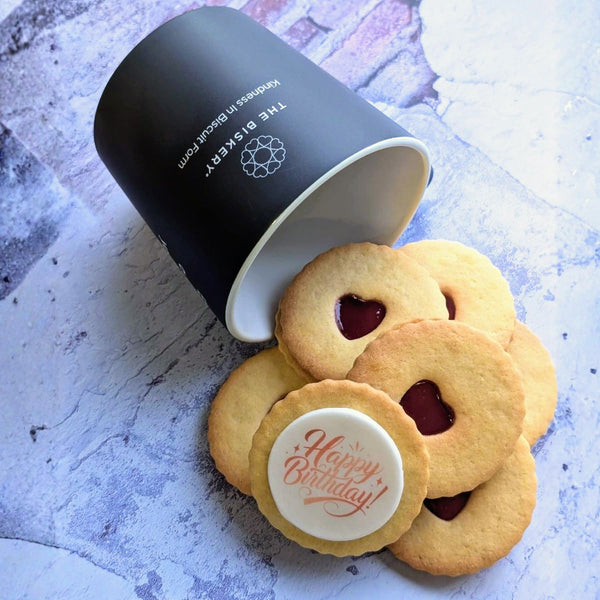 Jam Biscuits with a 'Happy Birthday' design on one of them. next to a black mug on a textured surface