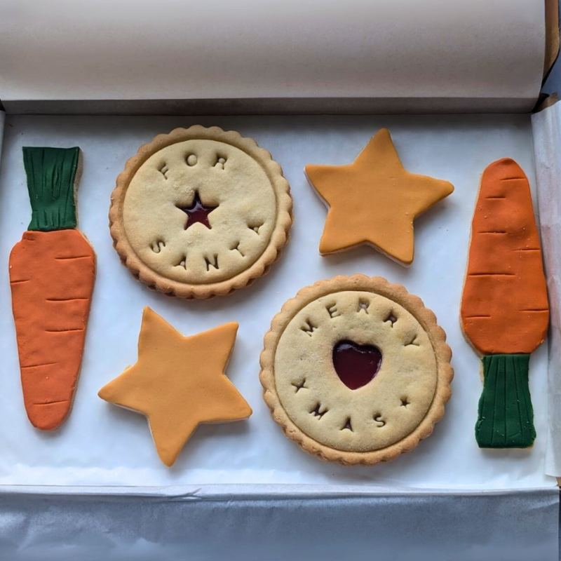 Cookies for Santa arranged in a festive flatlay, featuring star-shaped biscuits and carrot-shaped cookies on a white background, styled as a Christmas Eve cookie gift from The Biskery.