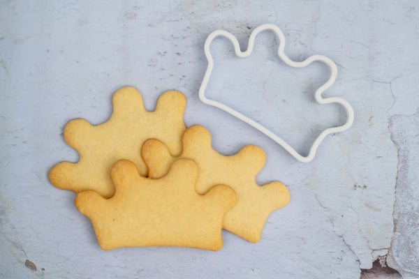 Cookie cutter in the shape of a crown. Next to 3 crown shaped biscuits that were made using the bespoke 3d printed cookie cutter.