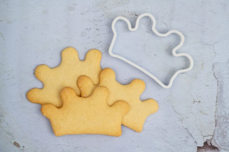 Cookie cutter in the shape of a crown. Next to 3 crown shaped biscuits that were made using the bespoke 3d printed cookie cutter.