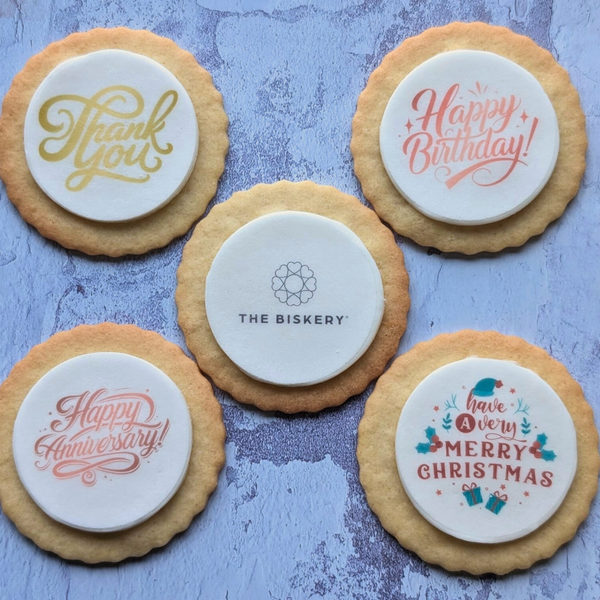 Biscuit with messages 'Thank You', 'Happy Birthday', 'The Biskery', 'Happy Anniversary', and 'Have a Very Merry Christmas' on a textured blue surface.