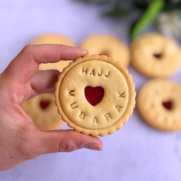 Hand holding a cookie with 'Hajj Mubarak' text and a heart shape, with more cookies in the background.