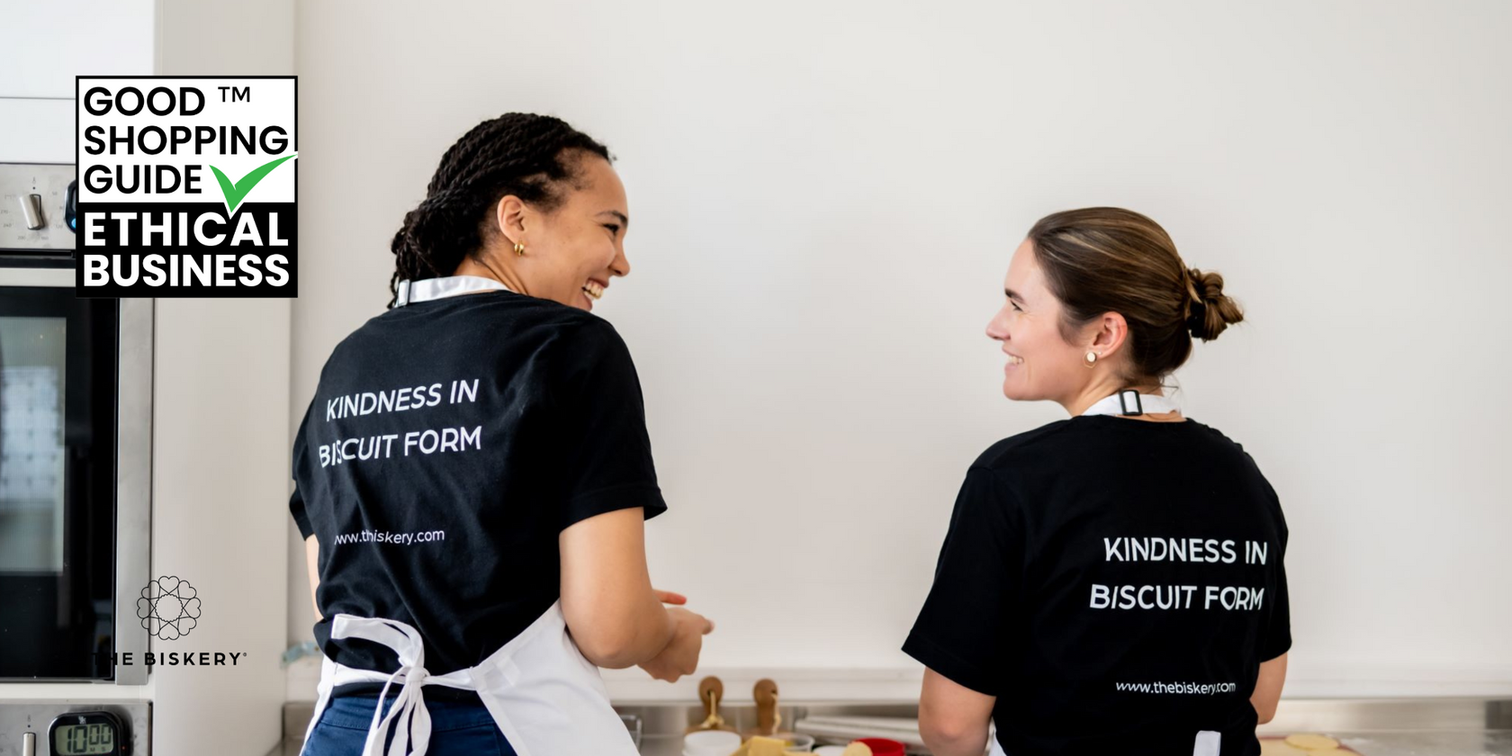 The Biskery co-founders in a kitchen wearing black t-shirts with 'Kindness in Biscuit Form' text, with a 'Good Shopping Guide Ethical Business' badge in the corner.