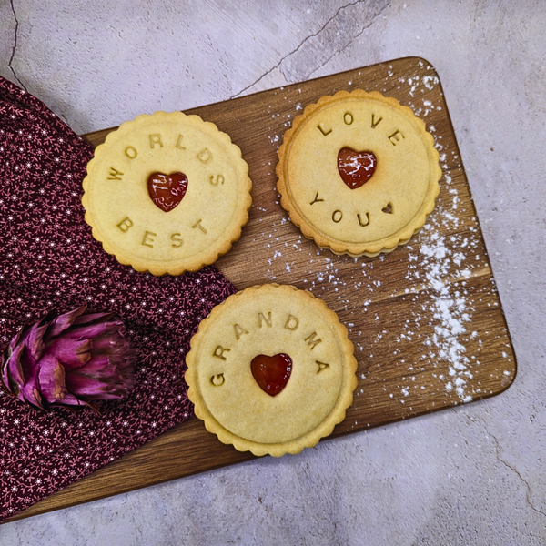 Three jam biscuits with messages 'Worlds Best', 'Love You', and 'Grandma' on a wooden board. Handmade in The Biskery.