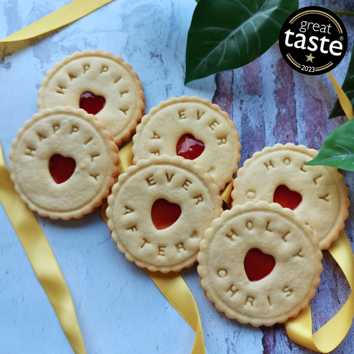 A selection of handcrafted Happily Ever After Biscuits with heart-shaped jam centres, personalised lettering, and a yellow ribbon on a styled background.