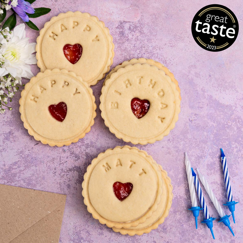 Happy birthday jam biscuits with heart-shaped cut-outs in the middle. Some of the biscuits are personalised with a name, and the background is purple