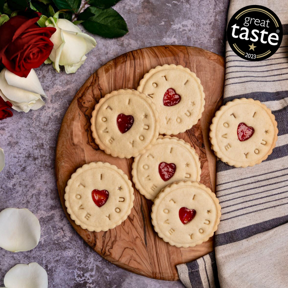 Wooden chopping board with heart-decorated biscuits saying 'I love you' and roses, next to personalised jam biscuits from The Biskery company