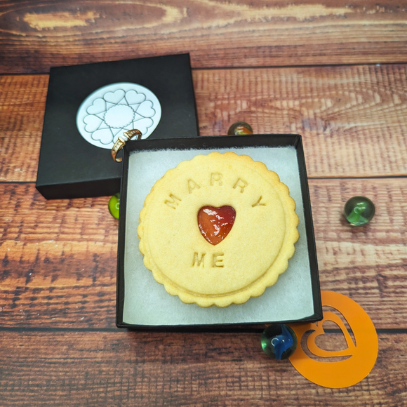 Biscuit in a box with the words "MARRY" and "ME". The biscuit has a heart shape cut out of it.
