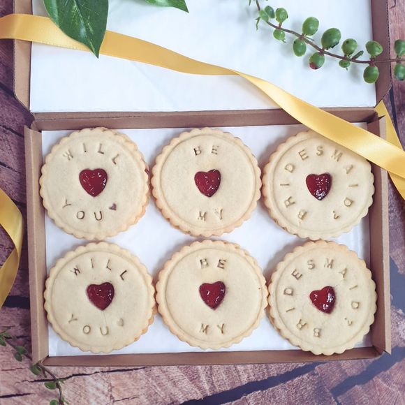 Neat row of six heart-shaped Jam biscuits in a box, a popular British treat with Red Currant jam. Each biscuit is decorated with "Will You Be My Bridesmaid?"