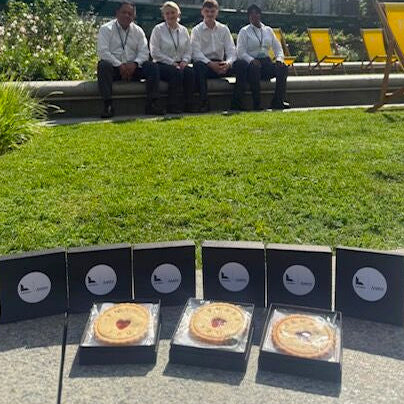 Four packaged jam biscuits displayed on a patio in front of a grassy lawn and modern apartment building, with four people sitting on a bench in the background.