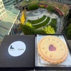 A jam biscuit with a heart-shaped cut out center and 'THANK YOU' embossed on top, presented in a clear compostable sleeve next to a black gift box with a white 'Aspire' logo, set against a background of a modern building courtyard.