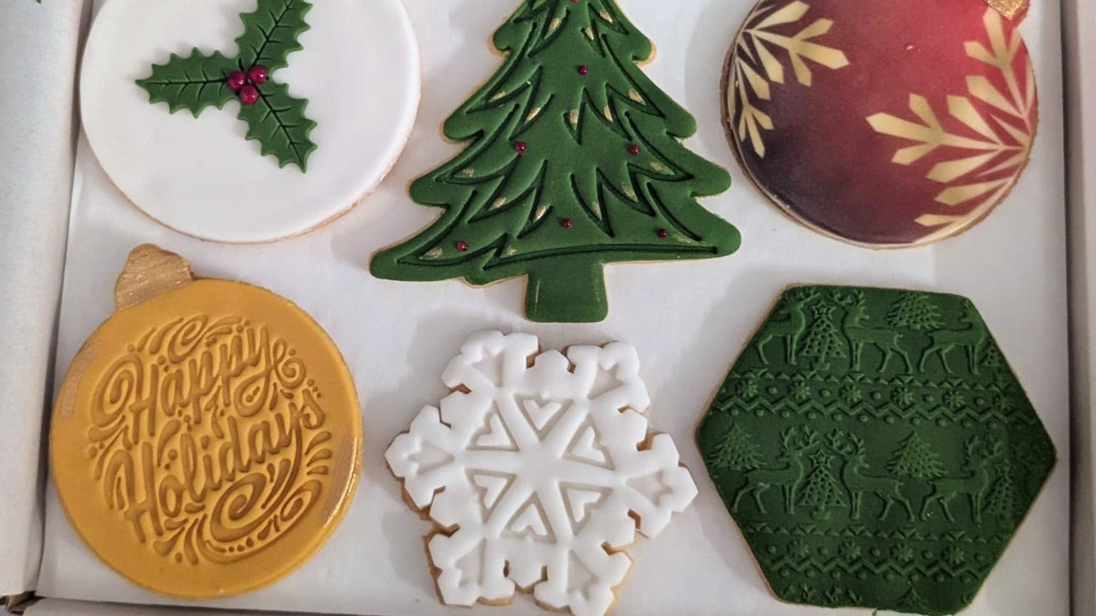 A boxed set of six decorated Christmas-themed cookies, including a holly cookie, a tree cookie, a red ornament cookie, a gold 'Merry Merry' ornament cookie, a white snowflake cookie, and a dark green hexagon cookie, displayed in a gift box with tissue paper and green leaves.