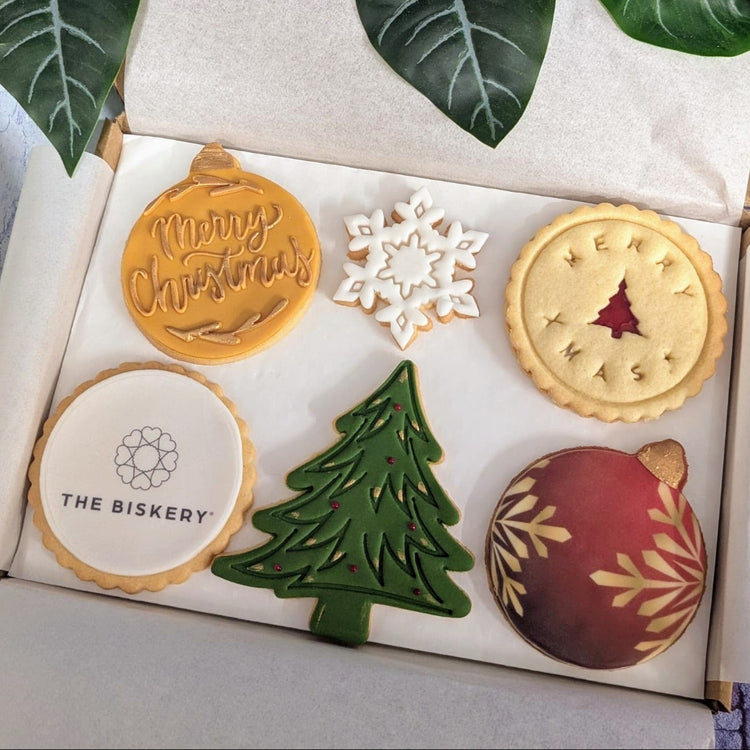 An open gift box containing six decorated Christmas bscuits, surrounded by green leaves. The biscuit selection include a gold 'Merry Christmas' ornament, a white snowflake, a jam biscuit wwith Christmas tree cut out, a white cookie that can ber personalised with your logo, a green Christmas tree, and a red bauble biscuit with gold details.