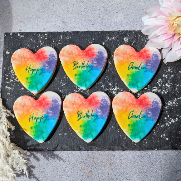 Six rainbow heart-shaped birthday cookies on a slate tray, personalised with messages like "Happy Birthday Charlie". 
