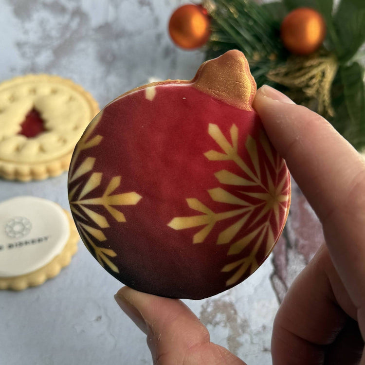 Red ornament biscuit with gold snowflake patterns held by a hand, with biscuits and Christmas decorations in the background.