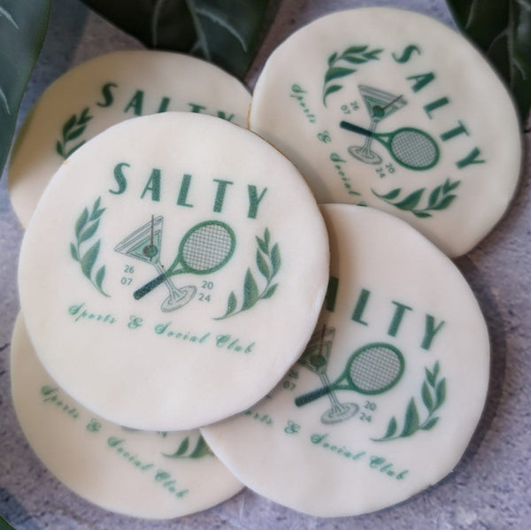 Five round branded biscuits with green printed tennis-themed graphics and the word 'SALTY' arranged on a textured gray surface with green leaves in the background.