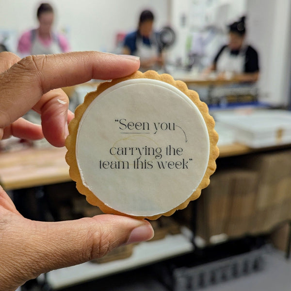 A hand holds a round, scalloped-edge cookie in a bakery setting. The center is white with the printed black text: 'Seen you carrying the team this week'