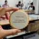 A hand holds a round, scalloped-edge cookie in a bakery setting. The center is white with the printed black text: 'Seen you carrying the team this week'