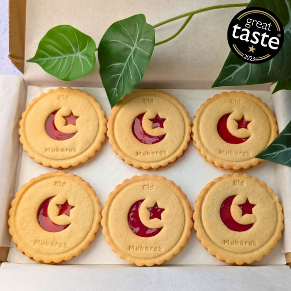 Six round, golden biscuits with red star and crescent jam centres are arranged in a white box. Each is printed with "Eid Mubarak". Green leaves are visible. Star and Crescent Eid Biscuits.
