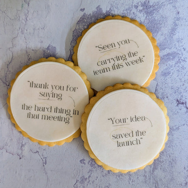Three round iced cookies with yellow scalloped edges and black printed motivational quotes, arranged on a textured light gray surface with green leaves in the corners.