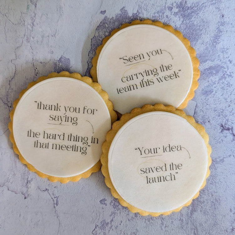 Three round iced cookies with yellow scalloped edges and black printed motivational quotes, arranged on a textured light gray surface with green leaves in the corners.