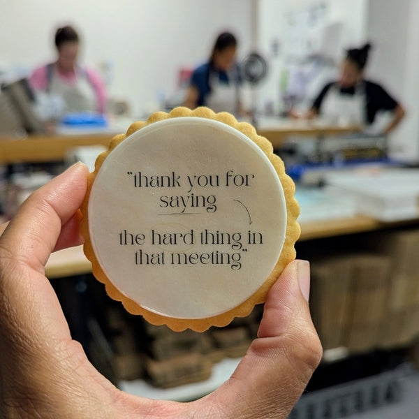 A hand holds a round, scalloped-edge cookie in a brightly lit bakery or workshop. The item has a pale cream center with black printed text that reads, "thank you for saying the hard thing in that meeting.