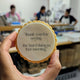 A hand holds a round, scalloped-edge cookie in a brightly lit bakery or workshop. The item has a pale cream center with black printed text that reads, "thank you for saying the hard thing in that meeting.