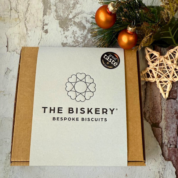 Boxes labelled 'The Biskery' and 'Bespoke biscuits' sit on a table, filled with Giant Merry Christmas Cookie. Blue sleeved biscuit box letterbox-friendly.
