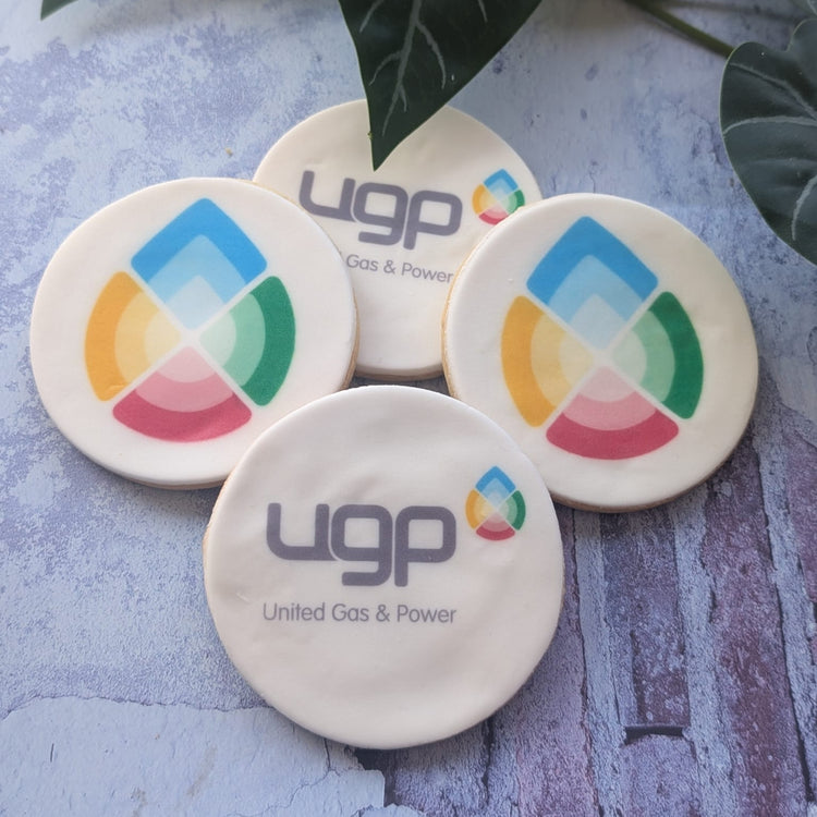 Four round branded biscuits featuring the UGP United Gas & Power logo and a colourful segmented circular graphic, arranged on a textured light blue surface with dark green leaves at the top.