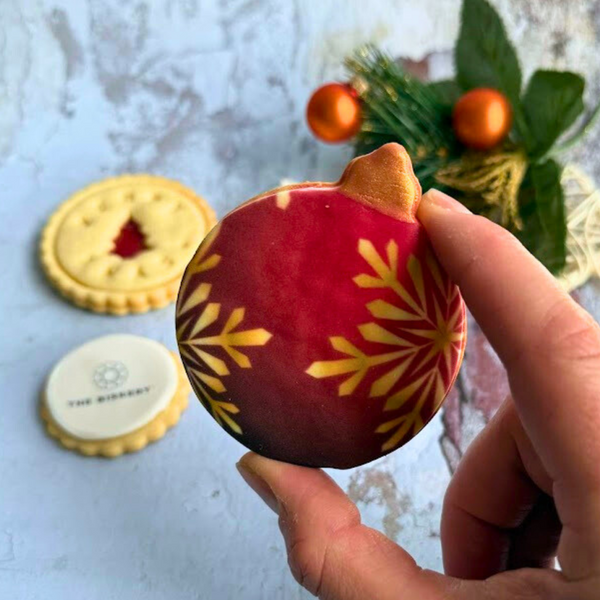 Hand holding a red and gold Christmas ornament with cookies and a tree in the background