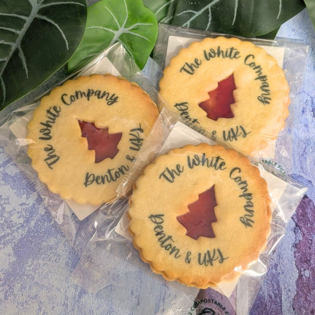 Three round baked cookies in clear compostable bags, each with a tree-shaped cutout showing a red center and the words 'The White Company' and 'Benton & UKL' written in black ink. The cookies are arranged on a textured light purple surface with green leaves in the background.