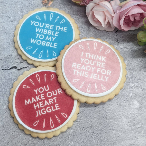 Three round sugar cookies decorated with colorful icing and white text, arranged on a gray stone surface with pink flowers in the background.