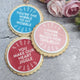 Three round sugar cookies decorated with colorful icing and white text, arranged on a gray stone surface with pink flowers in the background.