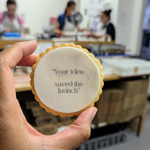 A hand holds a round scalloped-edge cookie in a bakery setting, with the center printed with the phrase “Your idea saved the launch” in black script.