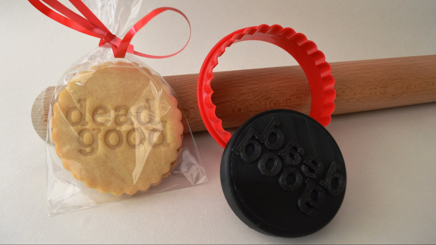 Cookie with 'dead good' text, wrapped in clear plastic with red ribbon, next to a black cookie stamp on a white background.