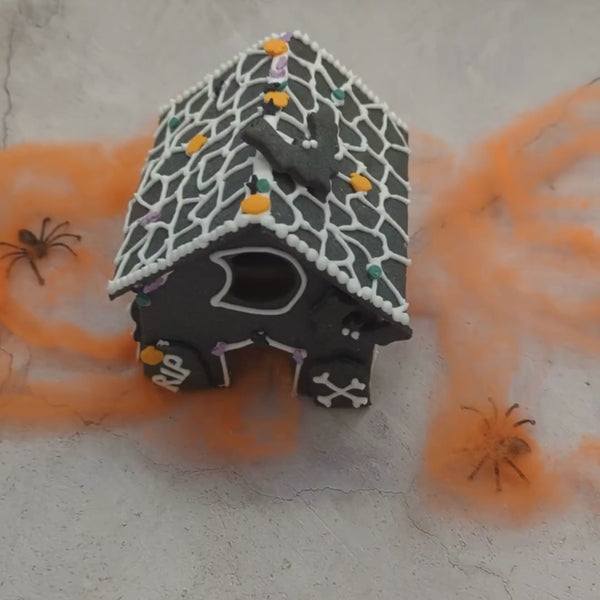 Haunted house decorating DIY kit. Small gingerbread house on a table, surrounded by spider webs, skeletons, and bats. Perfect for a spooky Halloween display.