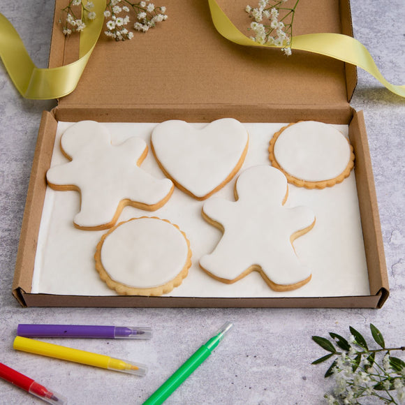 A variety of plain iced biscuits in letterbox friendly packaging on a purple background. The biscuits include a gingerbread man, heart, and circle biscuits, all decorated with white fondant.