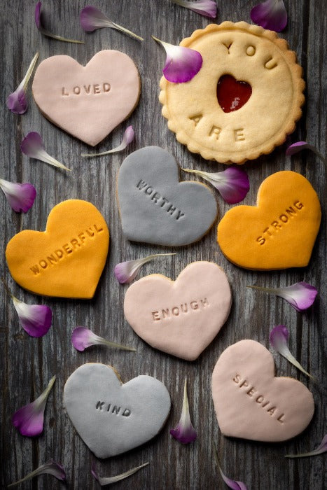 Affirmation biscuits from The Biskery. selection of heart-shaped biscuits. The biscuits are decorated with the words "LOVED", "WONDERFUL", "WORTHY", "ENOUGH", "STRONG", "KIND", and "SPECIAL" 