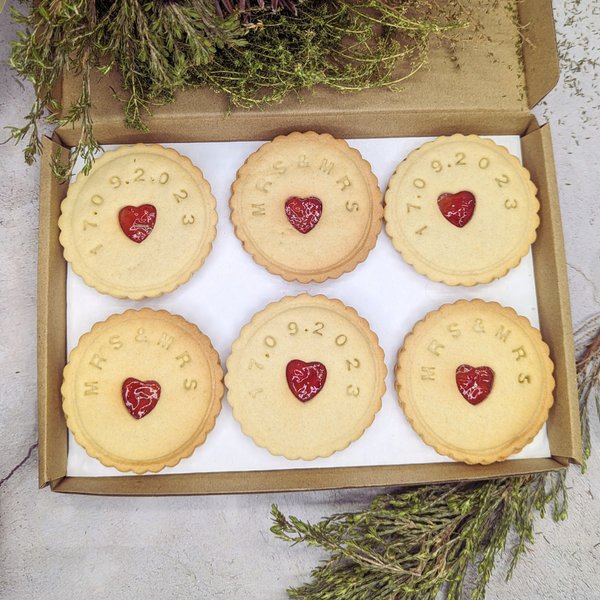 Box containing six Jam wedding biscuits, with heart-shaped biscuits at the centre, arranged on a table, adorned with a yellow ribbon for the MRS&MRS couple 